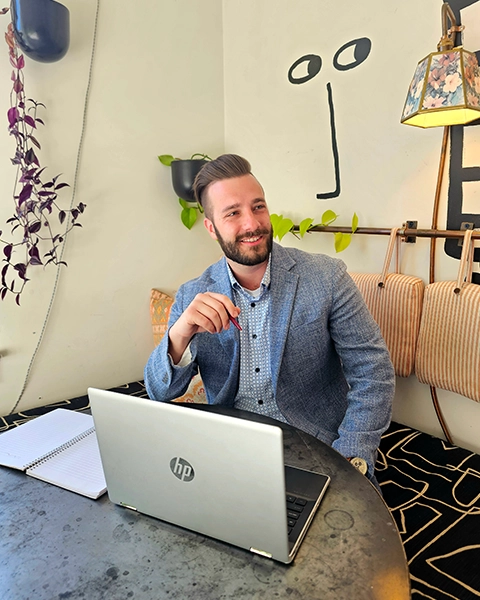 A person sits at a table with a laptop and notebook, surrounded by plants and a decorative lamp against a stylish wall.